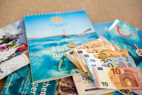Reisekataloge, Geldscheine, Münzen: Symbolbild fürs Sparen durch den Frühbucherrabatt