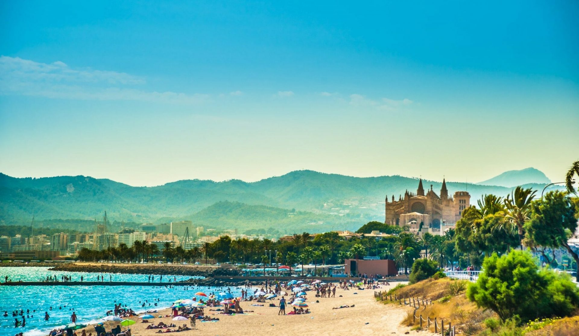 Urlaub Mallorca buchen bei REISEBÜRO Wache in Erfurt; im Bild: Mallorca, Vordergrund mit blauem Meer, Strand und Menschen, dahinter die Kathedrale von Palma de Mallorca, dahinter die Silhouette der mallorquinischen Berge