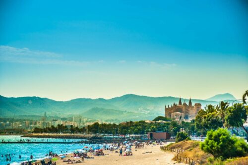 Urlaub Mallorca buchen bei REISEBÜRO Wache in Erfurt; im Bild: Mallorca, Vordergrund mit blauem Meer, Strand und Menschen, dahinter die Kathedrale von Palma de Mallorca, dahinter die Silhouette der mallorquinischen Berge