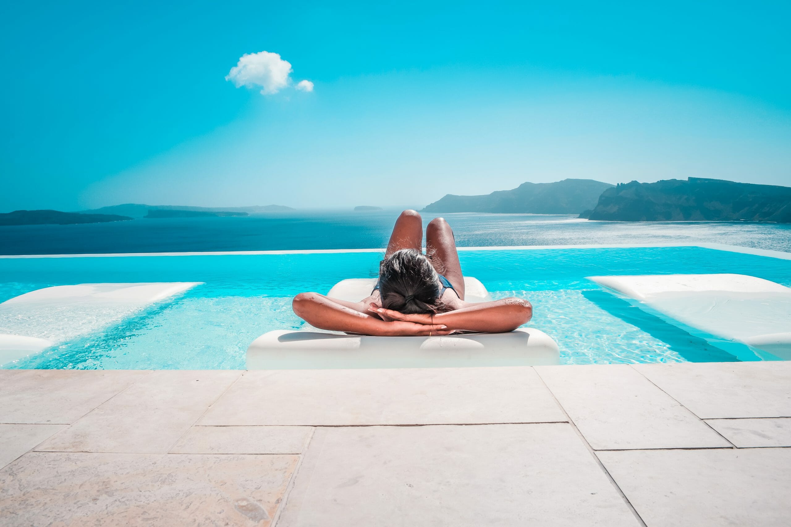 Erfurt REISEBÜRO Wache, Pauschalreise buchen: Symbolbild: Frau sonnt sich auf einer Liege im Infinity-Pool, im Hintergrund: das Meer und Berge