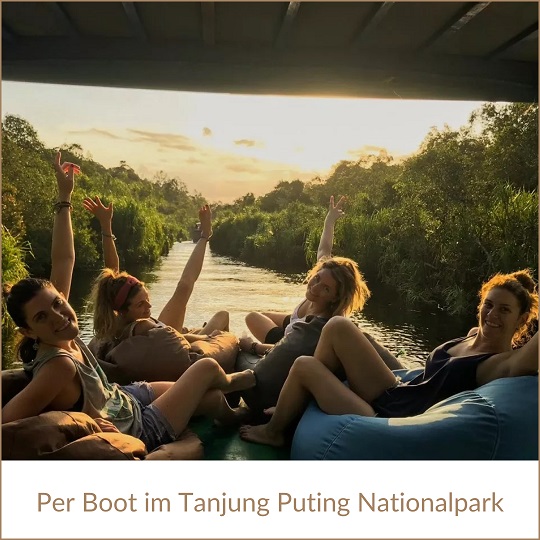 Borneo per Boot erkunden; im Bild der Blick aus einem Boot zu vier jüngere Frauen, die vor der geöffneten Luke auf Sitzsäcken sitzen und winken, dahinter der Fluss und rechts und links üppiges Grün des Tanjung Puting Nationalparks, die Sonne wirft goldenes Licht auf die Szene