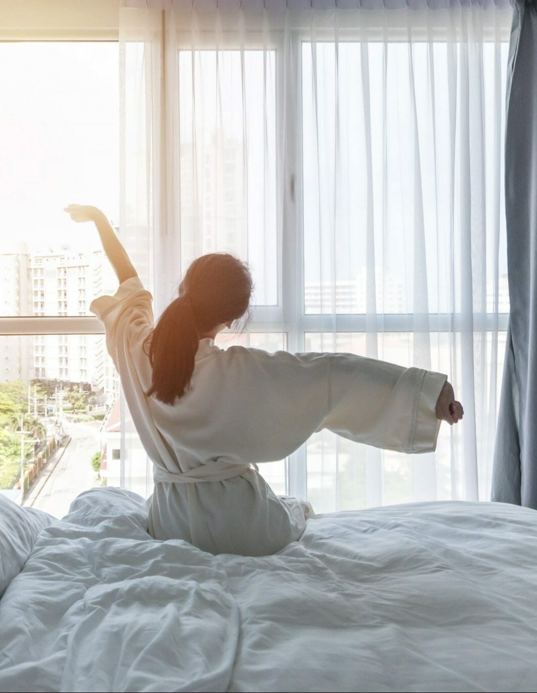 Erfurt REISEBÜRO Wache: Hotel buchen, Wellnesshotel buchen: Symbolbild: Frau im weißen Bademantel auf dem Bett vor einem großen Fenster, sie streckt sich