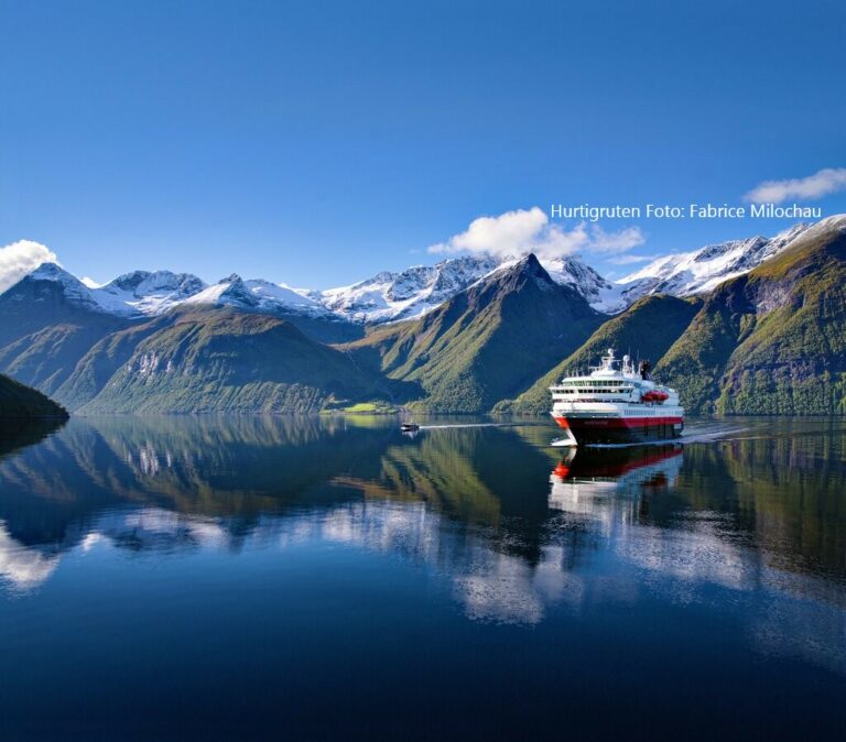 Hurtigruten buchen Erfurt bei REISEBÜRO Wache; im Bild: Postschiff im Hjörundfjord/Norwegen; im Hintergrund teils Schnee bedeckte, teils grüne Felsen, die sich im ruhigen Wasser spiegeln, das weiß-rot-schwarze Schiff fährt vor den Felsen