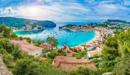 Urlaub Mallorca Balearen bei REISEBÜRO Wache buchen, im Bild: Port de Soller auf Mallorca, Blick von oben auf den Ort im Vordergrund mit Baumgrün, dahinter die türkisfarbene Bucht samt bewachsener Landzungen am Horizont