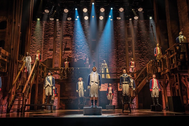 Hamilton, das Musical in Hamburg: Szenenbild bei dem Alexander Hamilton vorn auf einem Podest an der Bühne steht, im Hintergrund Soldaten