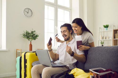 Erfurt REISEBÜRO Wache: Online-Check-In Flug, Symbolbild zeigt: einen Mann auf der Couch mit Laptop auf den Beinen, eine Frau mit Reisepass hinter ihm, beide lächeln, neben ihnen gelbe Koffer