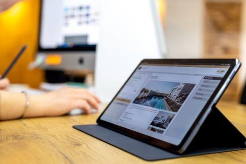 Erfurt REISEBÜRO Wache digitale Reiseanfrage senden, Symbolbild: aufgestelltes Tablet mit Ansicht der Reisebüro-Seite, im Hintergrund Arm und Computer einer Mitarbeiterin