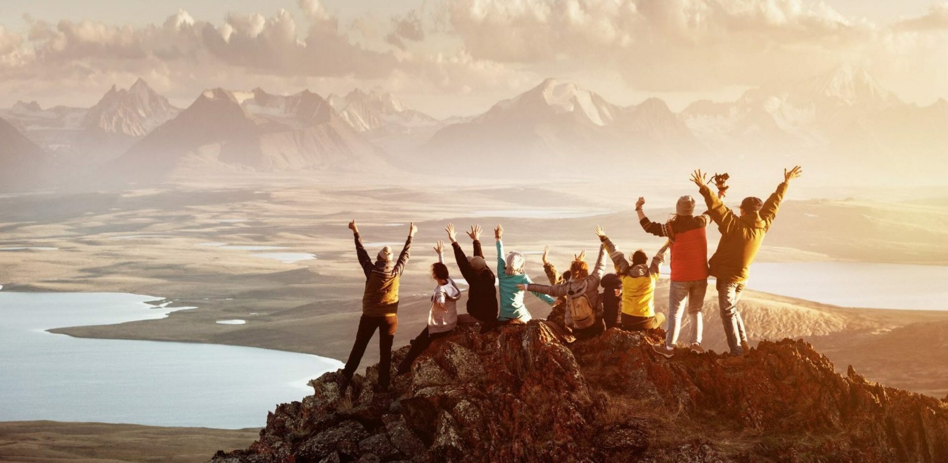 Erfurt REISEBÜRO Wache: Team, Wir über uns: Symbolbild: Menschen mit Rucksäcken, Jacken, Mützen, die die Arme in die Luft strecken auf einem Berg, im Hintergrund weitere Berge, alle von hinten von Sonne beschienen