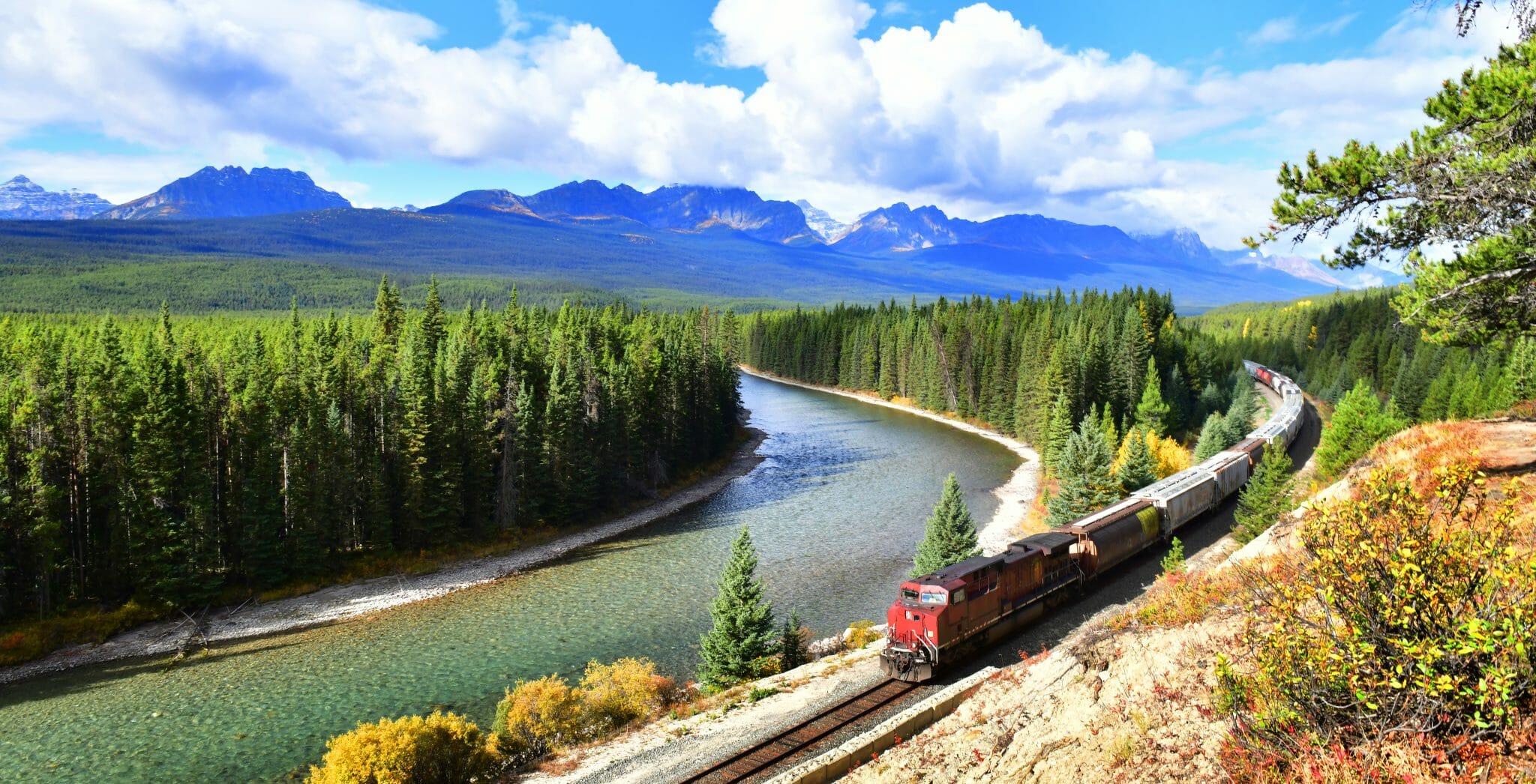 Kanada Rundreise buchen Erfurt bei REISEBÜRO Wache; im Bild: Zug fährt parallel zu einem Fluss im Banff-Nationalpark in Kanada, grüne Bäume, im Hintergrund die Silhouette der Berge