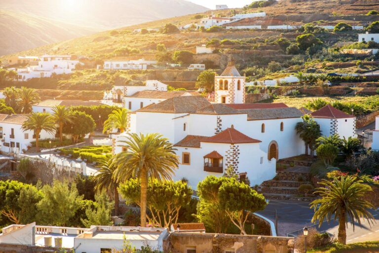 Kanaren buchen, zum Beispiel Urlaub auf Fuerteventura, bei REISEBÜRO Wache, Erfurt; im Bild: der Ort Betancuria, also weiße Häuser und eine weiße Kirche, eine Palme, im Hintergrund geht die Sonne unter