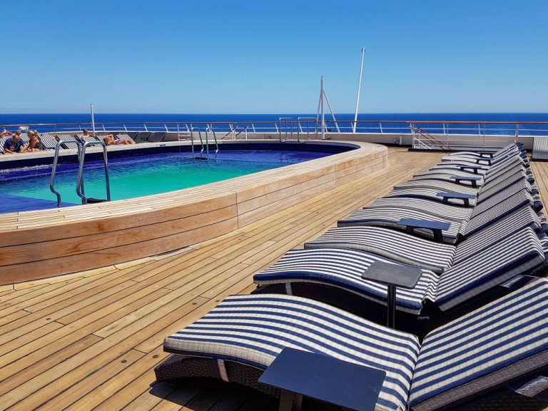 Erfurt Kreuzfahrt buchen von Nicko Cruises bei REISEBÜRO Wache; im Bild: Pooldeck auf der Vasco da Gama von Nicko Cruises, blauer, länglicher Pool, Sonnenliegen mit blau gestreiftem Bezug, Holzfußboden