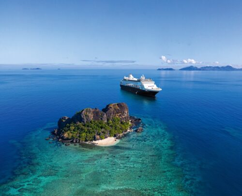 Kreuzfahrt Weltreise mit Nicko Cruises beim REISEBÜRO Wache buchen; im Bild: Meer mit kleiner, bewachsener Insel im Vordergrund, dahinter das Schiff Vasco da Gama von Nicko Cruises