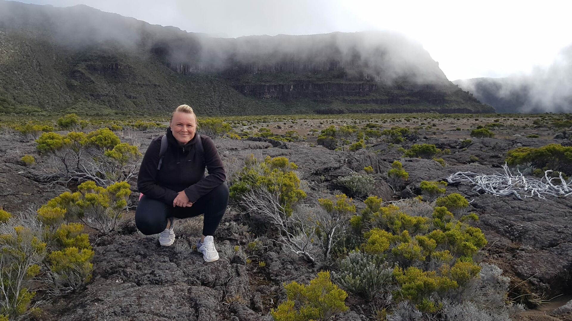 La Reunion Urlaub Aktivurlaub Buchen Erfurt Reisebuero Wache La Réunion Urlaub buchen Aktivurlaub buchen beim REISEBÜRO Wache in Erfurt; im Bild: Reiseverkehrskauffrau Anke Fröhlich, blond, in schwarzer Kleidung, weißen Turnschuhen, hockt auf einem erkalteten Lavafeld zwischen flachen grünen Pflanzen, im Hintergrund Berge in Rauchwolken