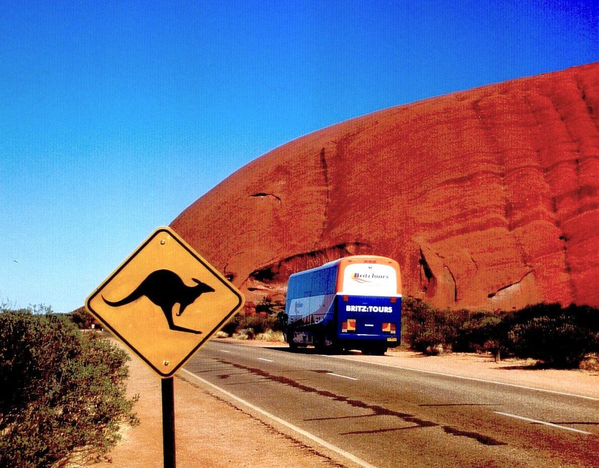 Meine Bucket List Reise nach Australien, zum Uluru; im Bild: Ein Straßenschild mit Känguru, ein Bus auf der Straße und im Hintergrund der rote heilige Berg Uluru bzw. Ayers Rock