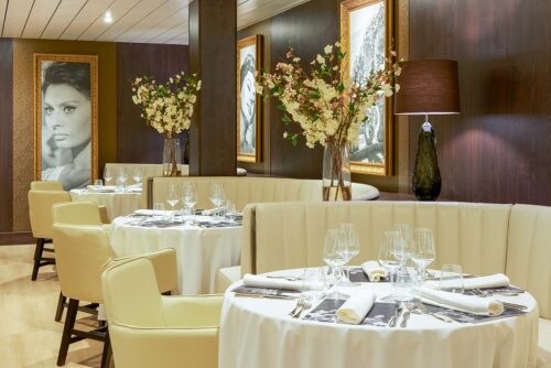 Restaurant von Nicko Cruises, Kreuzfahrt Weltreise buchen beim REISEBÜRO Wache, Erfurt; im Bild: mehrere eingedeckte Tische mit Weingläsern und weißem Porzellan, Blumen auf den runden Tischen, beigefarbene Möbel