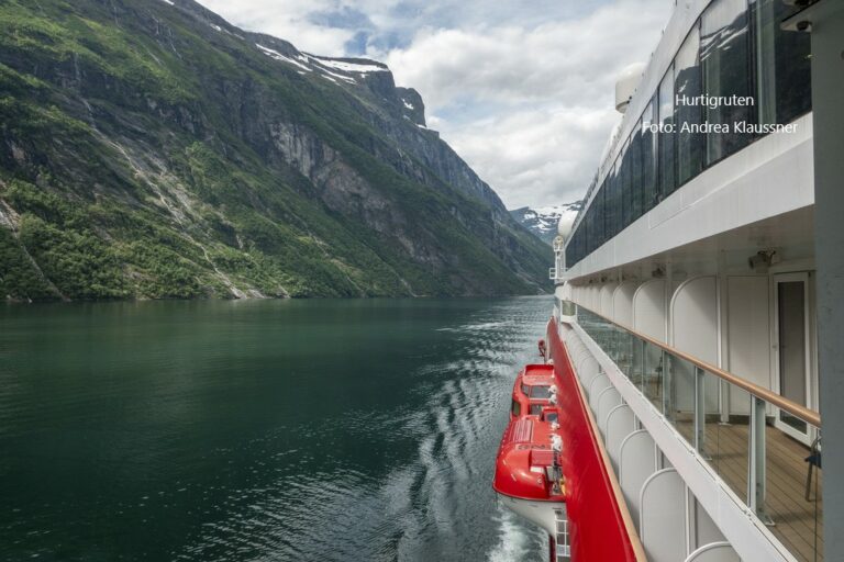 Postschiffroute Norwegen buchen Erfurt bei REISEBÜRO Wache; im Bild: links sind grünende Felsen, dann dunkles Meer, dann Balkone des Postschiffs von Hurtigruten in weiß ganz rechts