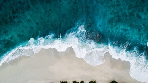 Blick von oben auf dunkeltürkisfarbene Meereswelle, die auf den Strand trifft - Symbolbild für eine digitale Reiseanfrage bei REISEBÜRO Wache