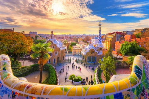 Urlaub Spanien buchen bei REISEBÜRO Wache Erfurt; im Bild: der Park Guell in Barcelona, Blick über den steinernen, bunten, geschwungenen Handlauf der Treppen in die Stadt hinunter, am Himmel scheint Sonne