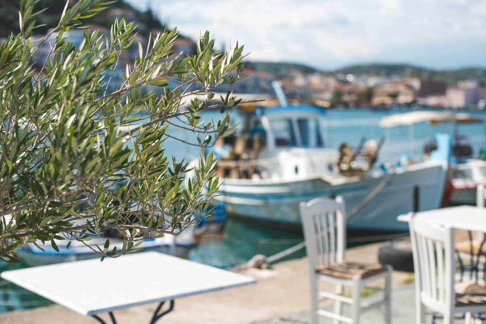 Griechenland Urlaub buchen bei REISEBÜRO Wache Erfurt; im Bild: Blick aus einem Restaurant auf Tische, Boot im Hafen, blaues Meer und im Hintergrund schroffe Berge