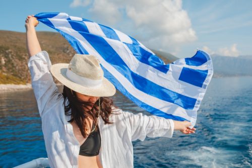 Symbolbild Griechenland 2023: Junge Frau hält die griechische Flagge wehend zwischen den Händen, im Hintergrund Meer und Berge