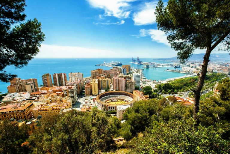 Spanien Urlaub buchen bei REISEBÜRO Wache Erfurt; im Bild: Blick auf Málaga, im Vordergrund Bäume und Büsche, dahinter, leicht abfallend Häuser und Hochhäuser, hinter ihnen blaues Meer und Hafen, blauer Himmel mit Wölkchen