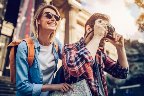 Symbolbild: Ausflug buchen für zu Hause, im Bild: junger Mann fotografiert, junge Frau mit Sonnenbrille lächelt