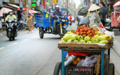 Vietnam Urlaub buchen bei REISEBÜRO Wache Erfurt; im Bild: Straßenszene in Ho-Chi-Minh-Stadt, im Vordergrund ein Straßenverkaufswagen mit frischem Obst, im Hintergrund Motorräder, Frau mit rundem, spitzen Hut